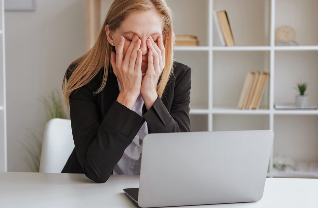 A woman sitting at a table in front of her laptop rubbing her eyes from eye strain.