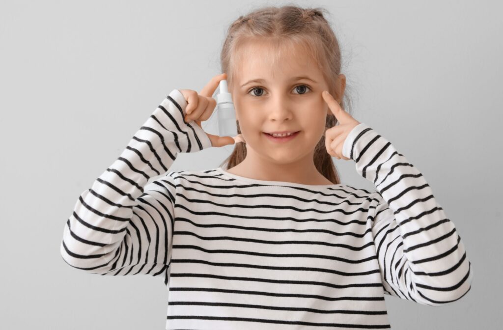 Young girl smiling while holding a bottle of eye drops, pointing to her eyes, wearing a striped shirt.