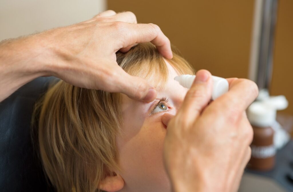 Adult gently administering eye drops to a young child seated in an optometrist office.