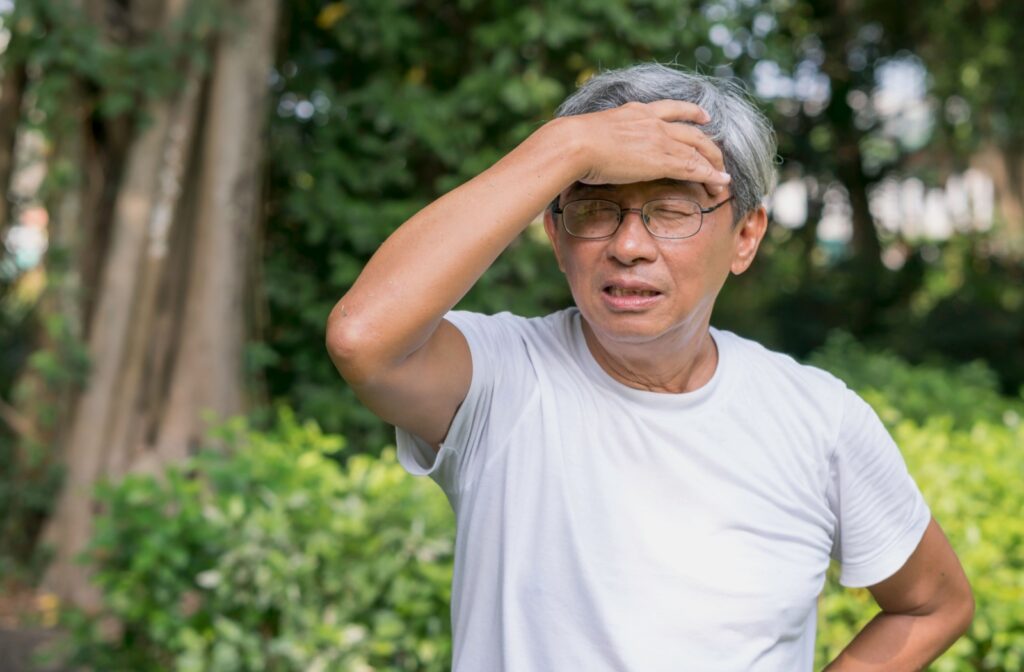 Older adult wearing glasses squinting and holding their forehead outdoors appearing distressed