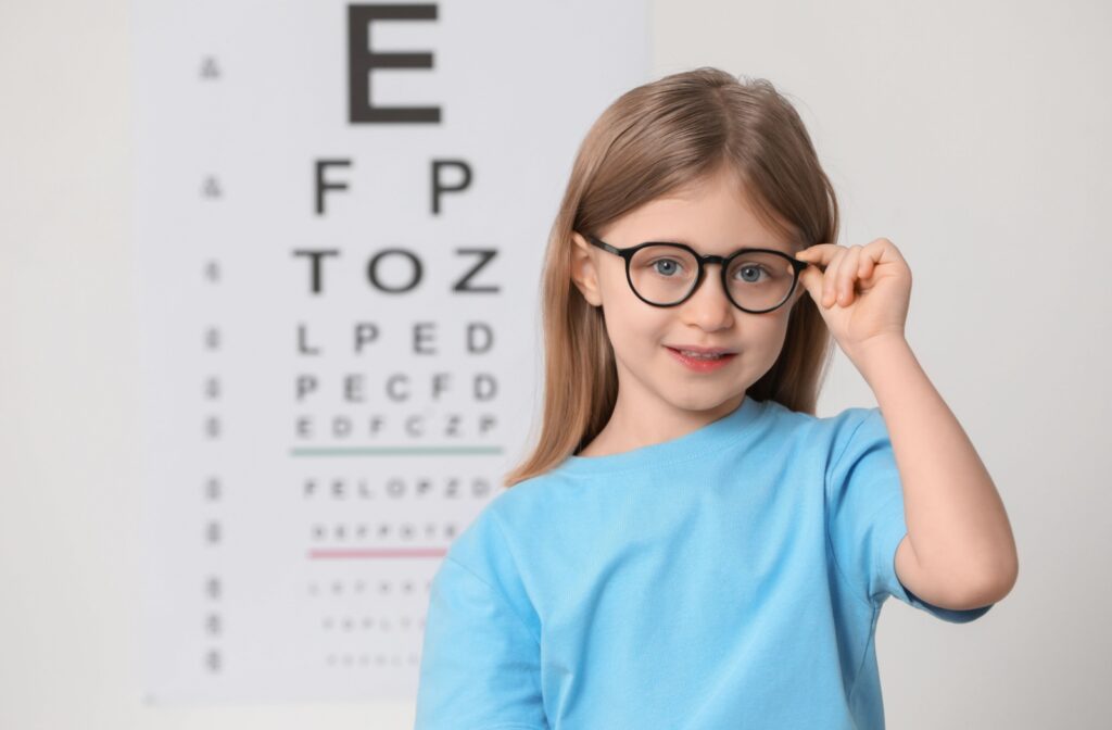 A child holds their glasses as they stand in front of a wall with a letter test for eye sight