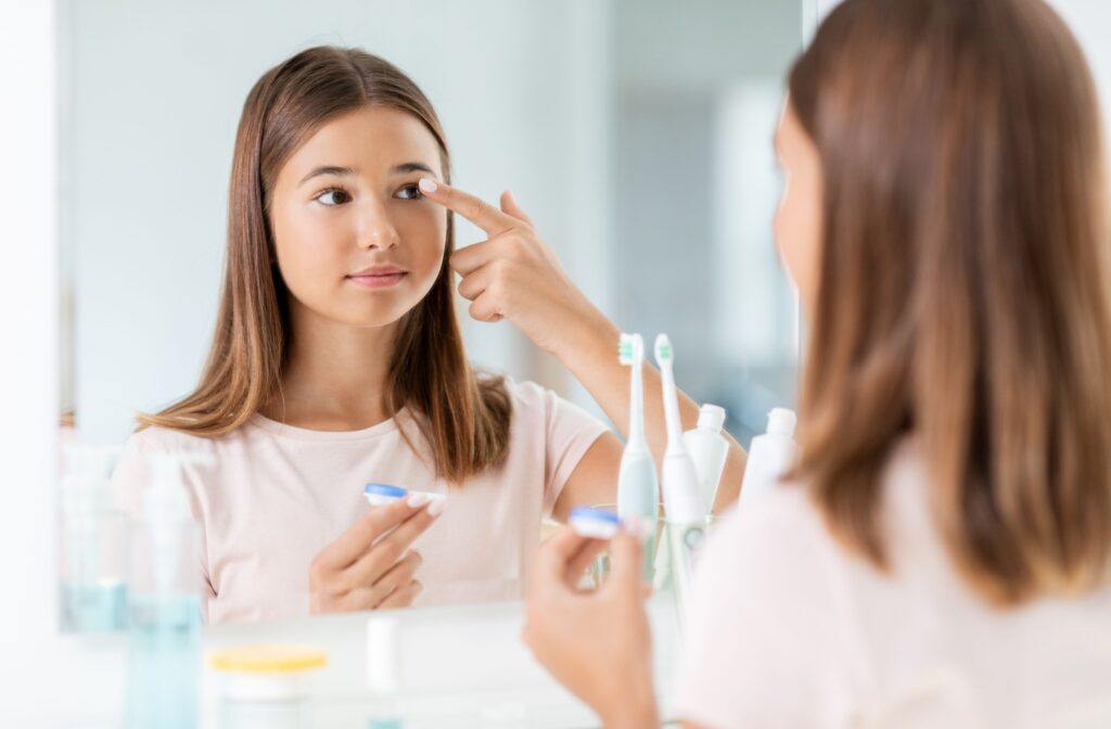 A teen holds a contact up to her eye while looking in the mirror